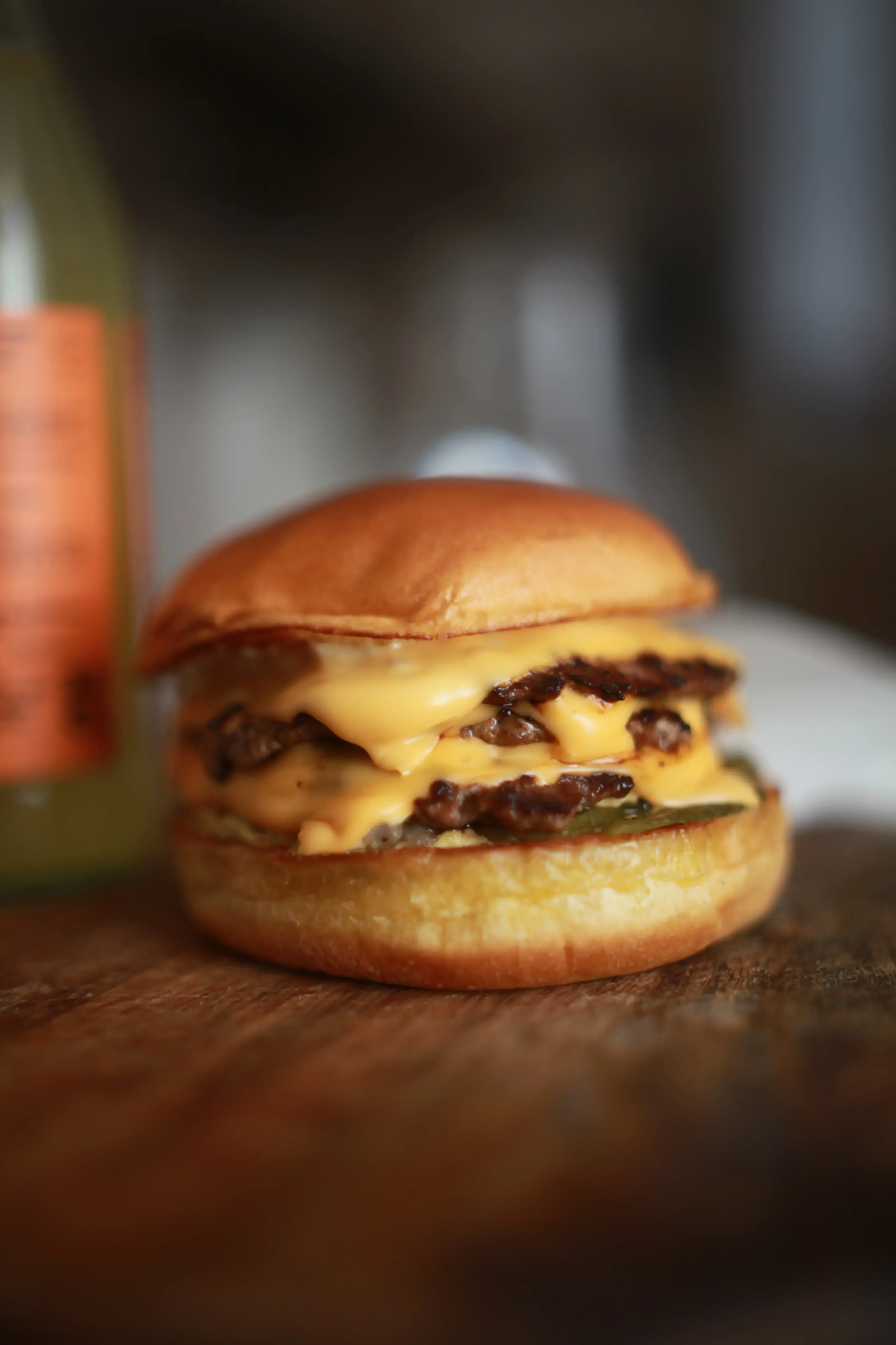 A shallow depth of field photo of a tripple smash burger next to a fruit kefir on a wooden serving board