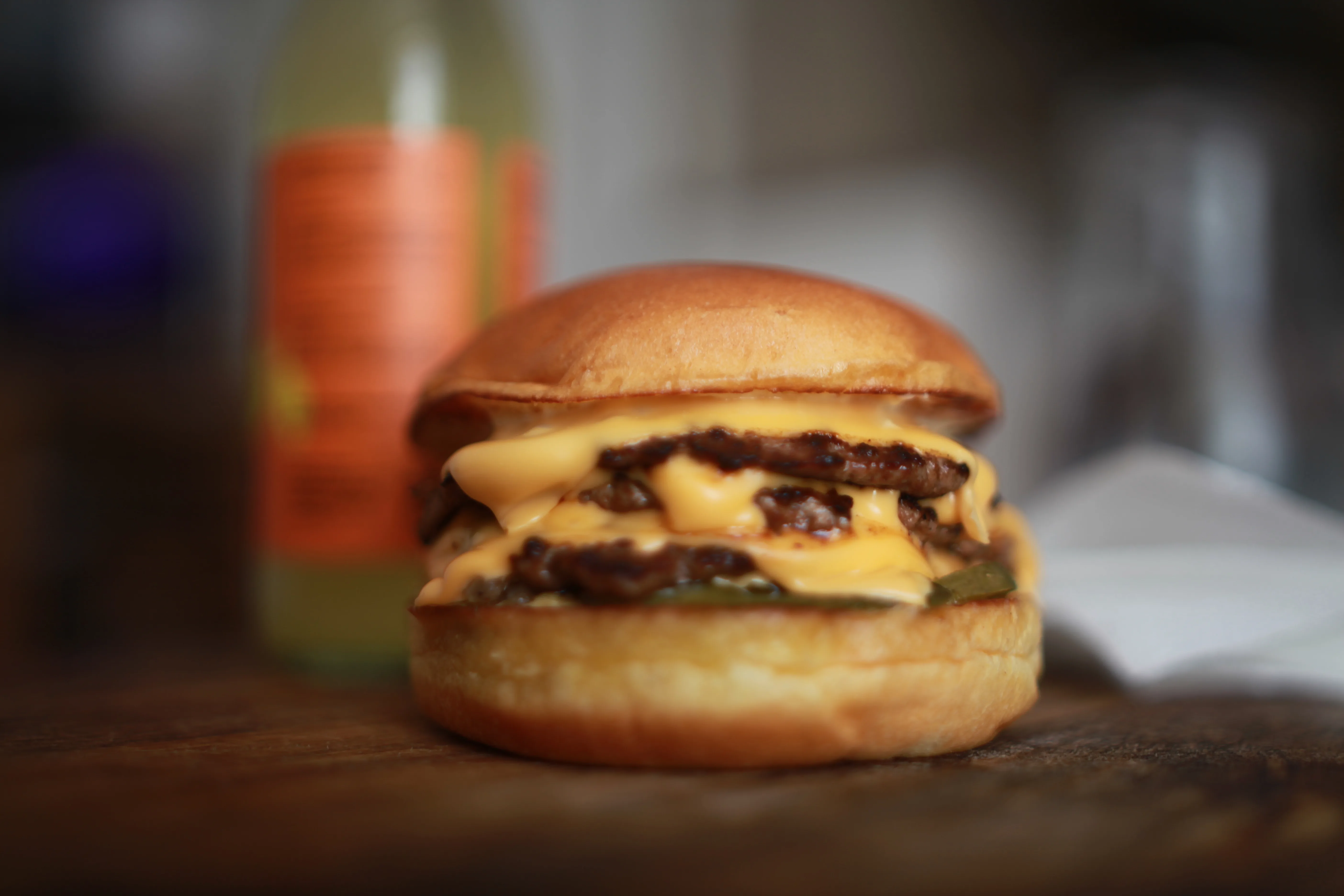 A shallow depth of field photo of a tripple smash burger next to a fruit kefir on a wooden serving board