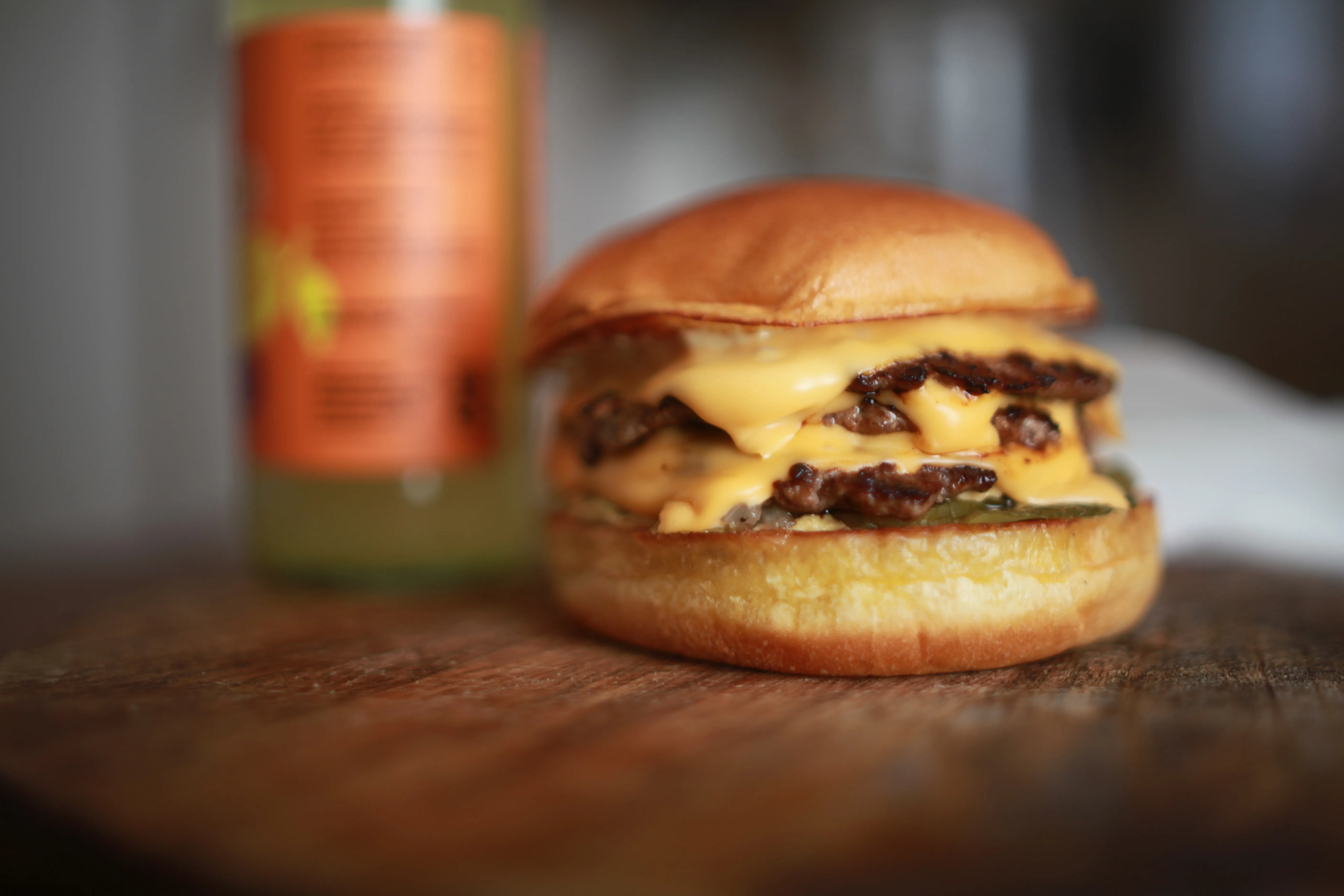 A shallow depth of field photo of a tripple smash burger next to a fruit kefir on a wooden serving board
